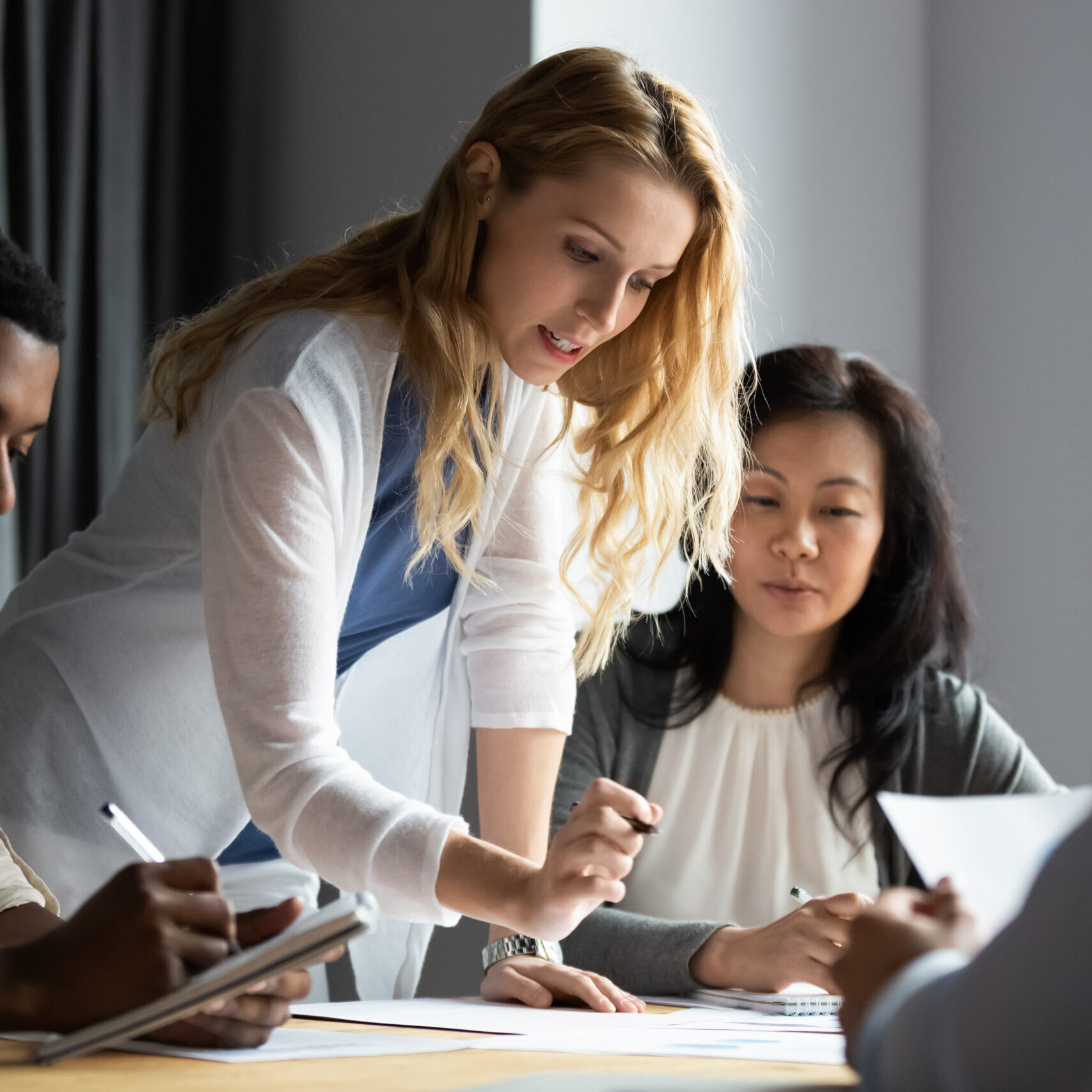 Serious millennial blonde female team leader analyzing financial or marketing paper document, reporting research results to focused multiracial young and older business partners colleagues at office.